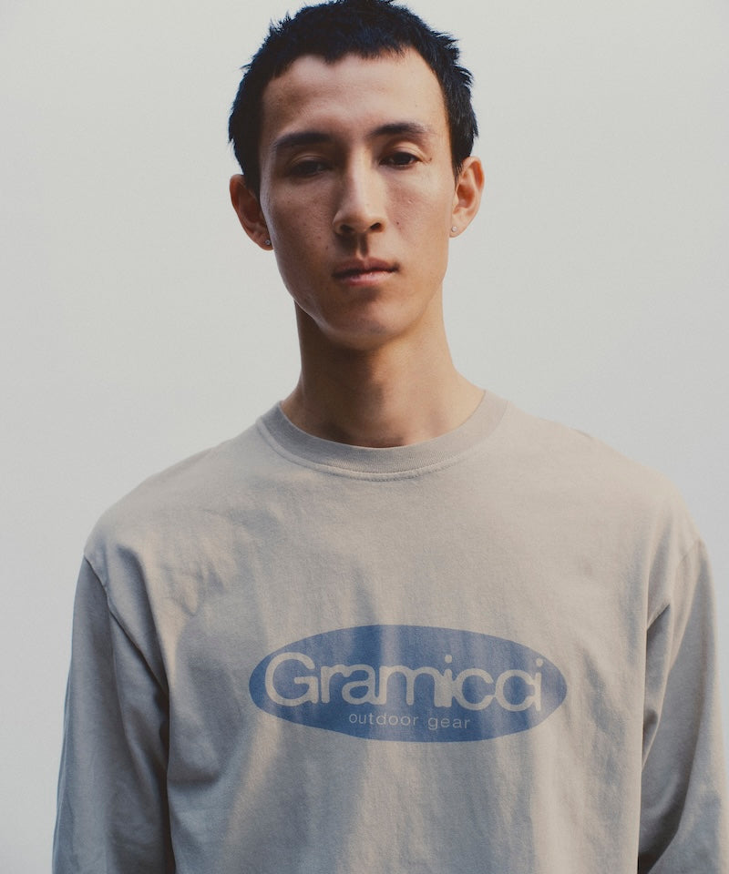 Model wearing a beige Gramicci Outdoor Gear L/S Tee with a blue graphic against a light-tone background.