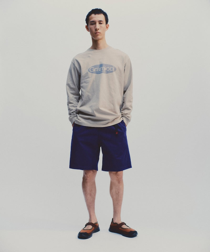 Model wearing a beige Gramicci Outdoor Gear L/S Tee with a blue graphic and blue shorts against a light-tone background.