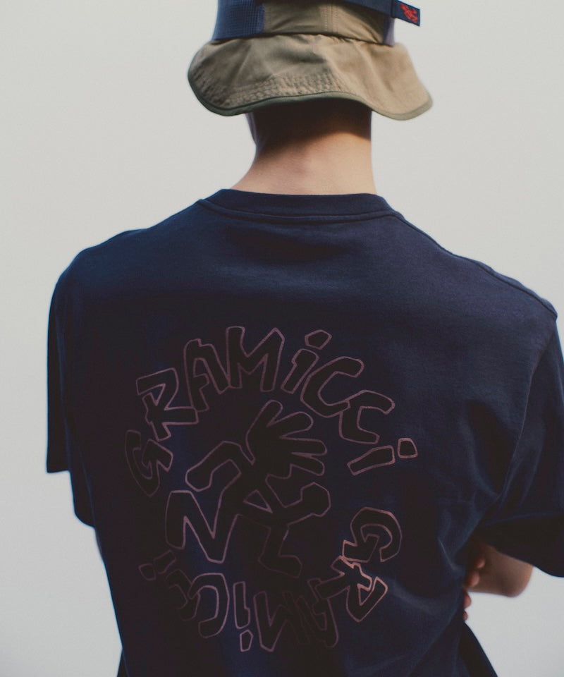 Rear view of a model wearing a navy Gramicci Running Man Tee with graphic against a light tone background.