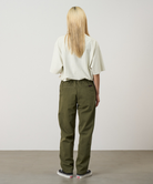 Back view of a model wearing olive green pants, a cream short-sleeve top, and black low-top sneakers, standing against a plain light gray studio background.