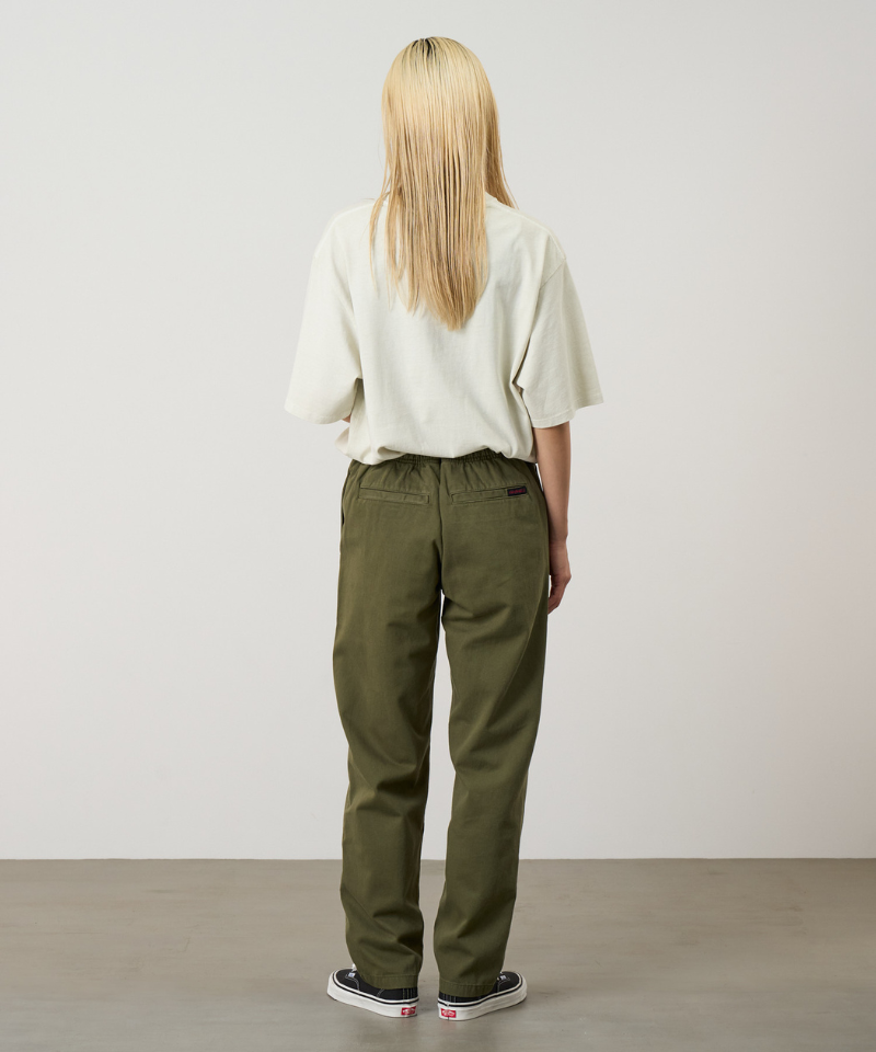Back view of a model wearing olive green pants, a cream short-sleeve top, and black low-top sneakers, standing against a plain light gray studio background.