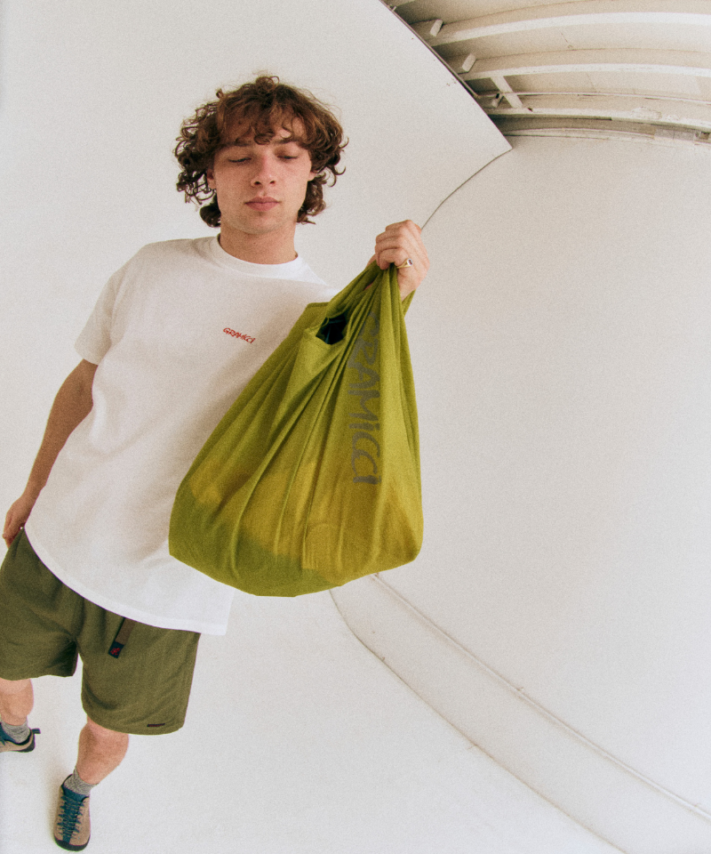 Model wearing a white shirt and green shorts holding an olive Gramicci Ultra-Light Shopper on a light-tone background.