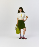 Model wearing a white shirt and a green skirt holding an olive Gramicci Ultra-Light Shopper on a light-tone background.