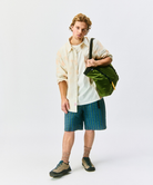 Model carying an olive Gamicci Trail Tote and wearing an orange shirt and blue plaid shorts on a light-tone background.