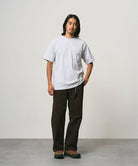 Model wearing a ash heather Gramicci One Point Tee and brown pants on a light-tone background.