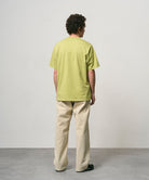 Back view of a model wearing a sweet pea colored Gramicci One Point Tee and beige pants on a light-tone background.