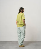 Back view of a model wearing a sweet pea colored Gramicci One Point Tee and light green pants on a light-tone background.