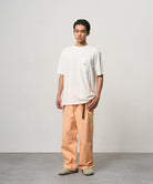 Model wearing a white Gramicci One Point Tee and orange pants on a light-tone background.