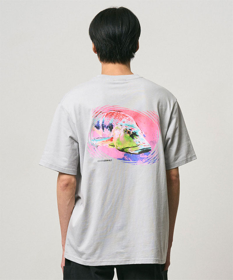 Back view of a model wearing a Gramicci mist colored Bass Tee with a graphic against a light-tone background.