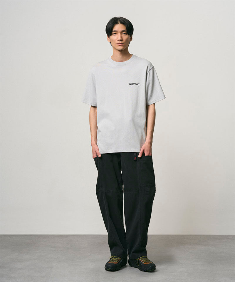 Model wearing a mist colored Gramicci Bass Tee and black pants on a light-tone background.