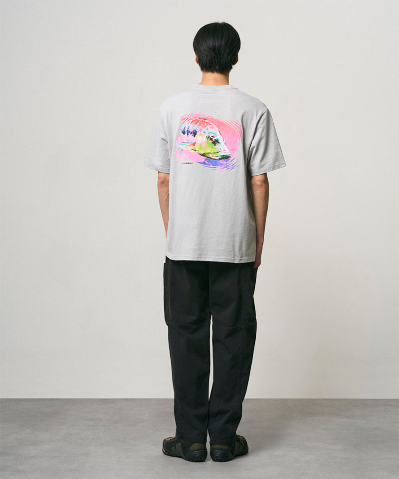 Back view of a model wearing a mist colored Gramicci Bass Tee with a graphic and black pants on a light-tone background.