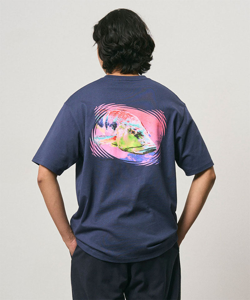 Back view of a model wearing a Gramicci navy Bass Tee with a graphic against a light-tone background.