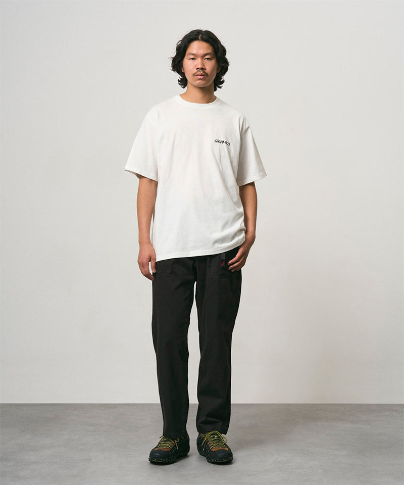 Model wearing a white Gramicci Bass Tee and black pants on a light-tone background.