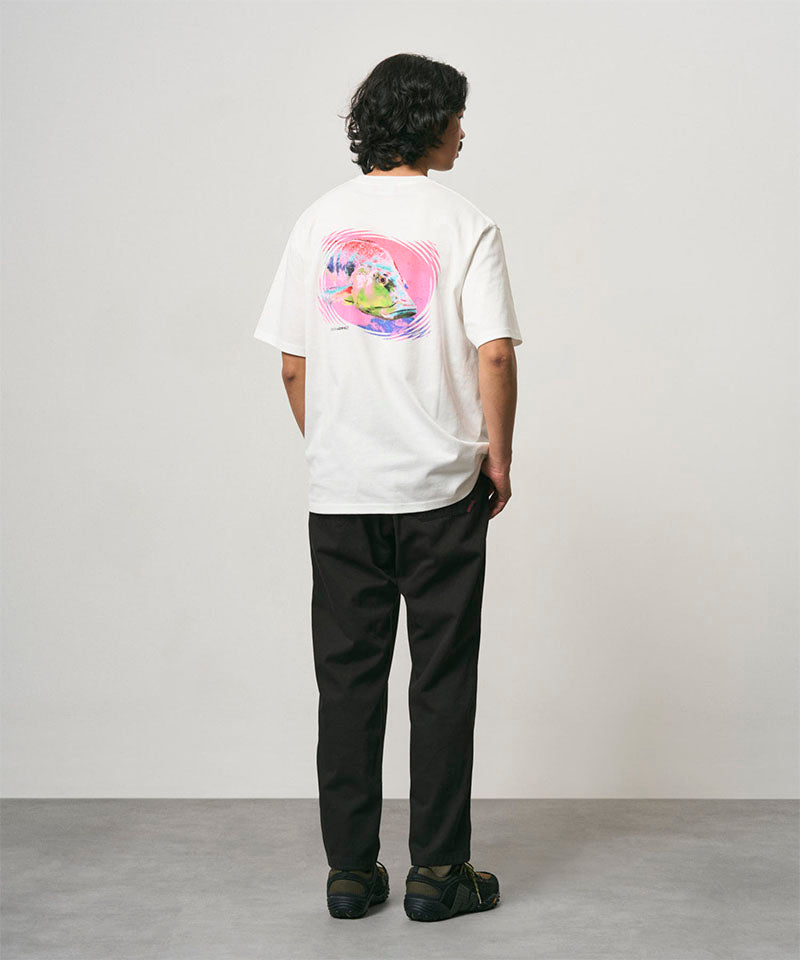 Back view of a model wearing a white Gramicci Bass Tee with a graphic and black pants on a light-tone background.