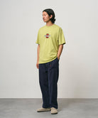 Model wearing a sweet pea colored Gramicci Ladybug Tee with colorful graphic and blue pants on a light-tone background.
