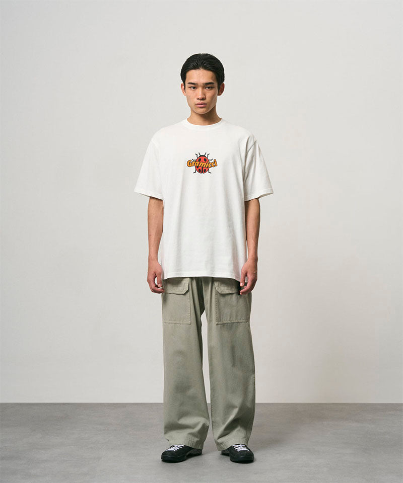 Model wearing a white Gramicci Ladybug Tee with colorful graphic and light green pants on a light-tone background.