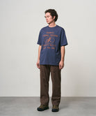 Model wearing a navy Gramicci Yosemite Valley Tee with a graphic and brown pants on a light-tone background.