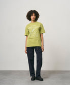 Model wearing a sweet pea colored Gramicci Yosemite Valley Tee with a graphic and black pants on a light-tone background.