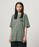 Model wearing a Gramicci sage pigment colored GCE Tee Pigment Dyed against a light-tone background.