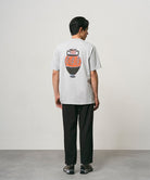 Back view of a model wearing a mist colored Gramicci Vase Tee with a graphic and black pants on a light-tone background.