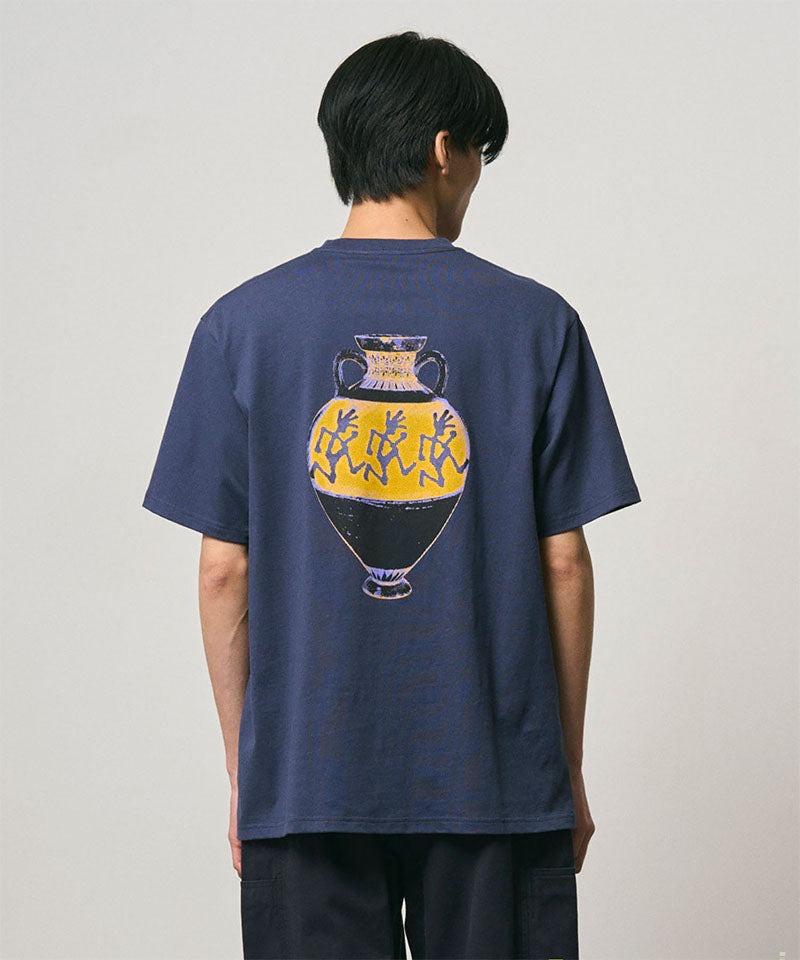 Back view of a model wearing a Gramicci navy Vase Tee with a graphic against a light-tone background.