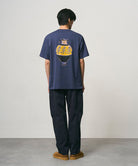Back view of a model wearing a navy Gramicci Vase Tee with a graphic and black pants on a light-tone background.