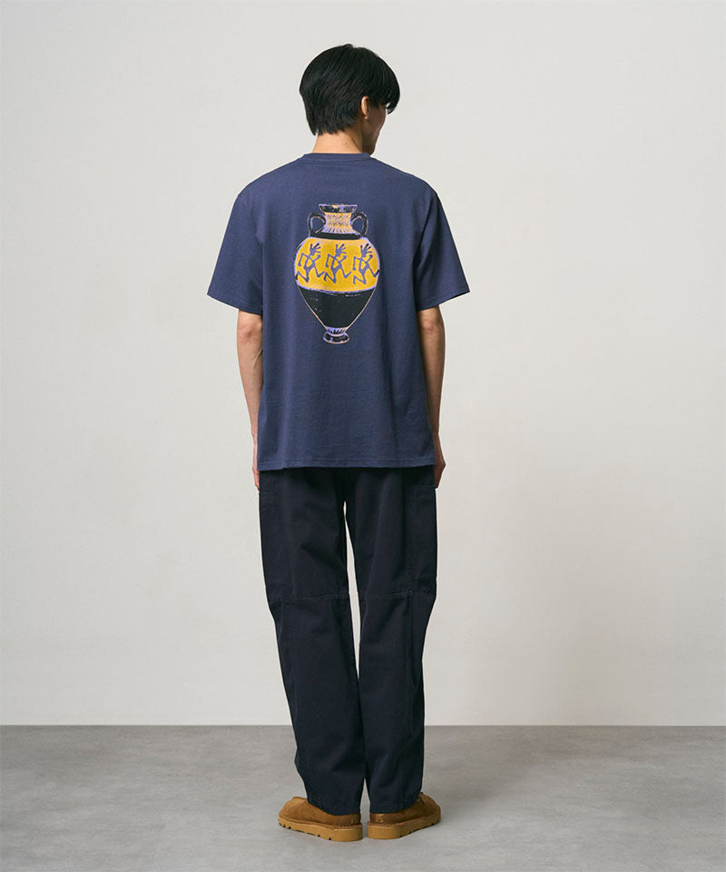 Back view of a model wearing a navy Gramicci Vase Tee with a graphic and black pants on a light-tone background.