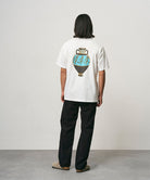 Back view of a model wearing a white Gramicci Vase Tee with a graphic and black pants on a light-tone background.