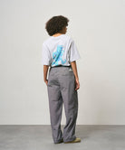 Back view of a model wearing a ash heather color Gramicci Joker Tee with a teal graphic and grey pants on a light-tone background.
