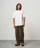 Model wearing a white Gramicci Joker Tee and light green pants on a light-tone background.