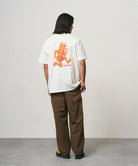 Back view of a model wearing a white Gramicci Joker Tee with an orange graphic and light green pants on a light-tone background.