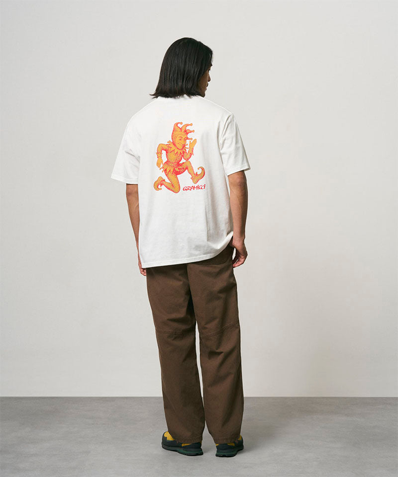 Back view of a model wearing a white Gramicci Joker Tee with an orange graphic and light green pants on a light-tone background.