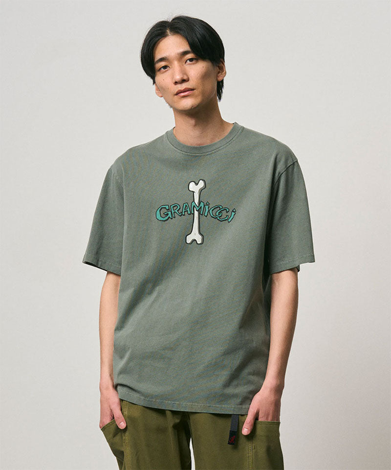 Model wearing a Gramicci sage pigment colored Bone Tee Pigment Dyed with a graphic against a light-tone background.