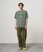 Model wearing a sage pigment colored Gramicci Bone Tee Pigment Dyed with a graphic and olive pants on a light-tone background.