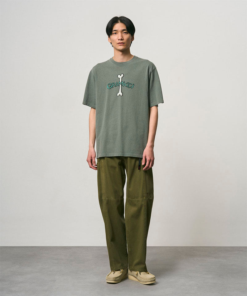 Model wearing a sage pigment colored Gramicci Bone Tee Pigment Dyed with a graphic and olive pants on a light-tone background.