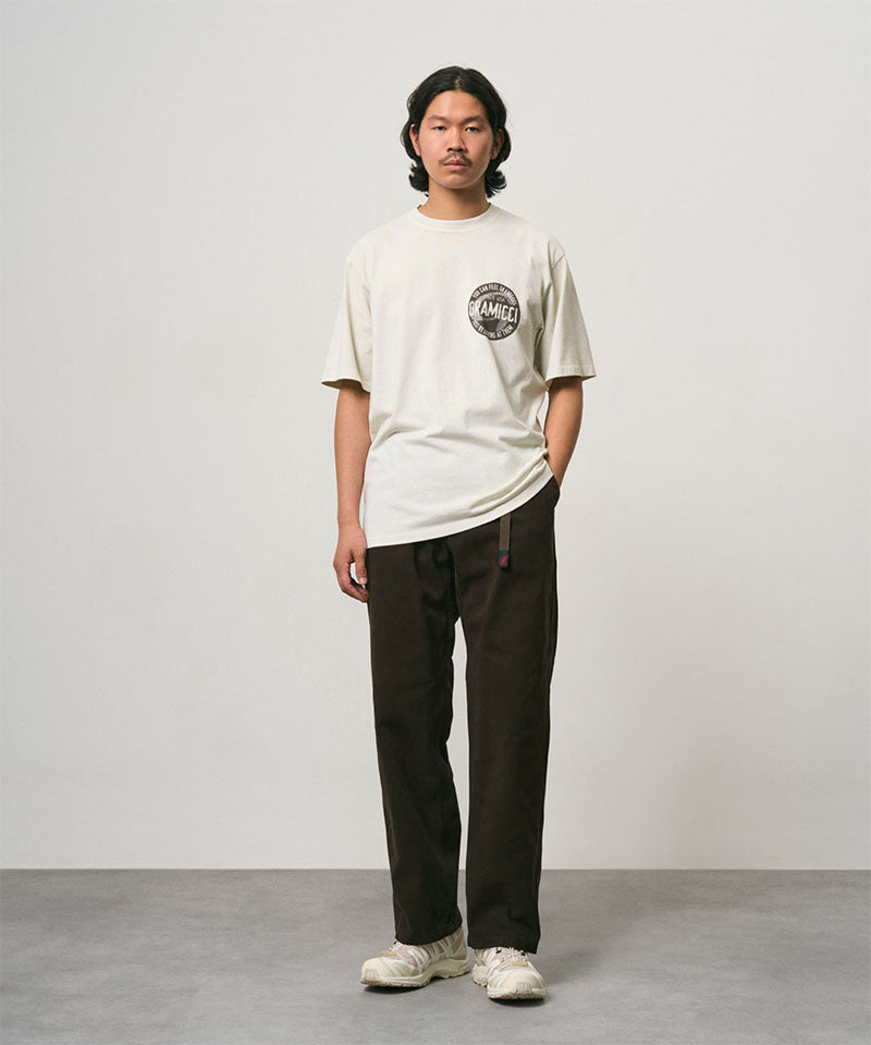Model wearing a natural pigment colored Gramicci Icon Tee Pigment Dyed with a graphic and dark brown pants on a light-tone background.