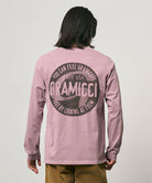 Back view of a model wearing a Gramicci lilac pigment colored Icon Long Sleeve Tee Pigment Dyed with a graphic against a light-tone background.