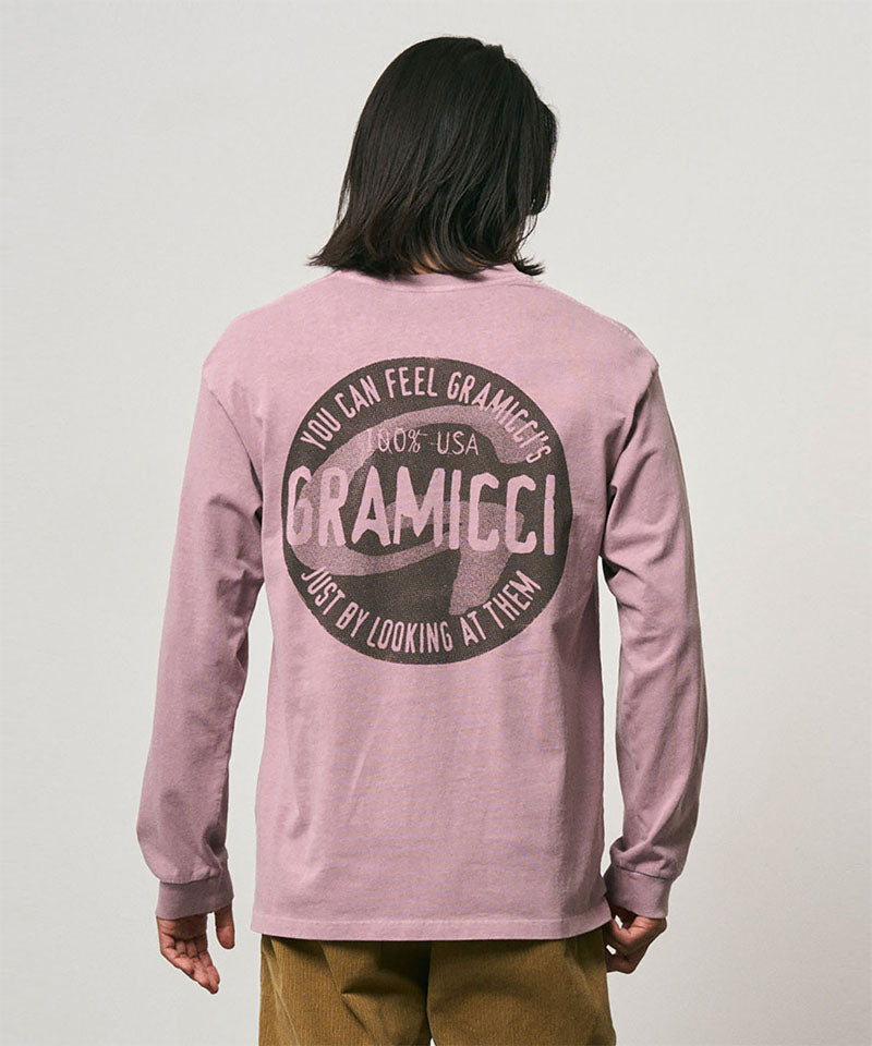 Back view of a model wearing a Gramicci lilac pigment colored Icon Long Sleeve Tee Pigment Dyed with a graphic against a light-tone background.