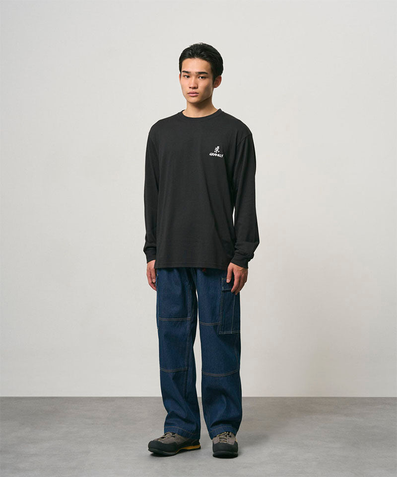 Model wearing a black Gramicci One Point Logo Long Sleeve Tee with a graphic and blue pants on a light-tone background.