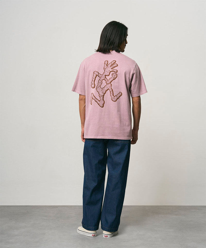 Back view of a model wearing a lillac pigment colored Gramicci Livin' Tee Pigment Dyed with a running man graphic and blue pants on a light-tone background.