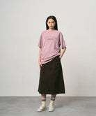 Model wearing a lillac pigment colored Gramicci Livin' Tee Pigment Dyed with a Livin' Under A Rock graphic and black skirt on a light-tone background.
