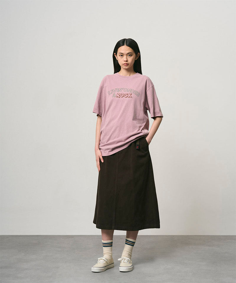 Model wearing a lillac pigment colored Gramicci Livin' Tee Pigment Dyed with a Livin' Under A Rock graphic and black skirt on a light-tone background.