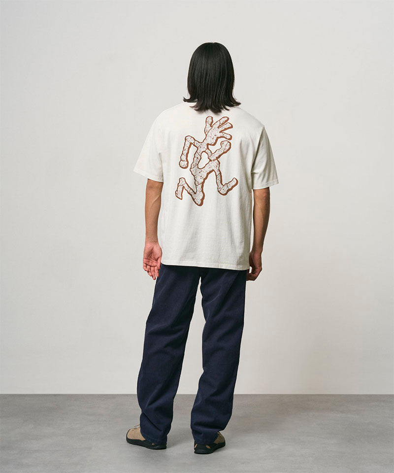Back view of a model wearing a natural pigment colored Gramicci Livin' Tee Pigment Dyed with a running man graphic and blue pants on a light-tone background.