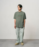 Model wearing a sage pigment colored Gramicci Livin' Tee Pigment Dyed with a Livin' Under A Rock graphic and light green pants on a light-tone background.