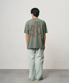 Back view of a model wearing a sage pigment colored Gramicci Livin' Tee Pigment Dyed with a running man graphic and light colored pants on a light-tone background.