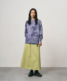 Model wearing a elm pigment colored Gramicci 5-Pocket Skirt and a purple tie-dye sweatshirt on a light-tone background.