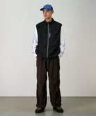Model wearing a black Gramicci Flyweight Vest and brown pants on a light-tone background.