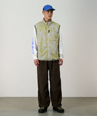 Model wearing a yellow and grey grid camo Gramicci Flyweight Vest and brown pants on a light-tone background.