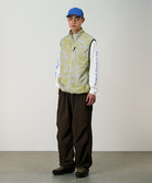 Model wearing a yellow and grey grid camo Gramicci Flyweight Vest and brown pants on a light-tone background.