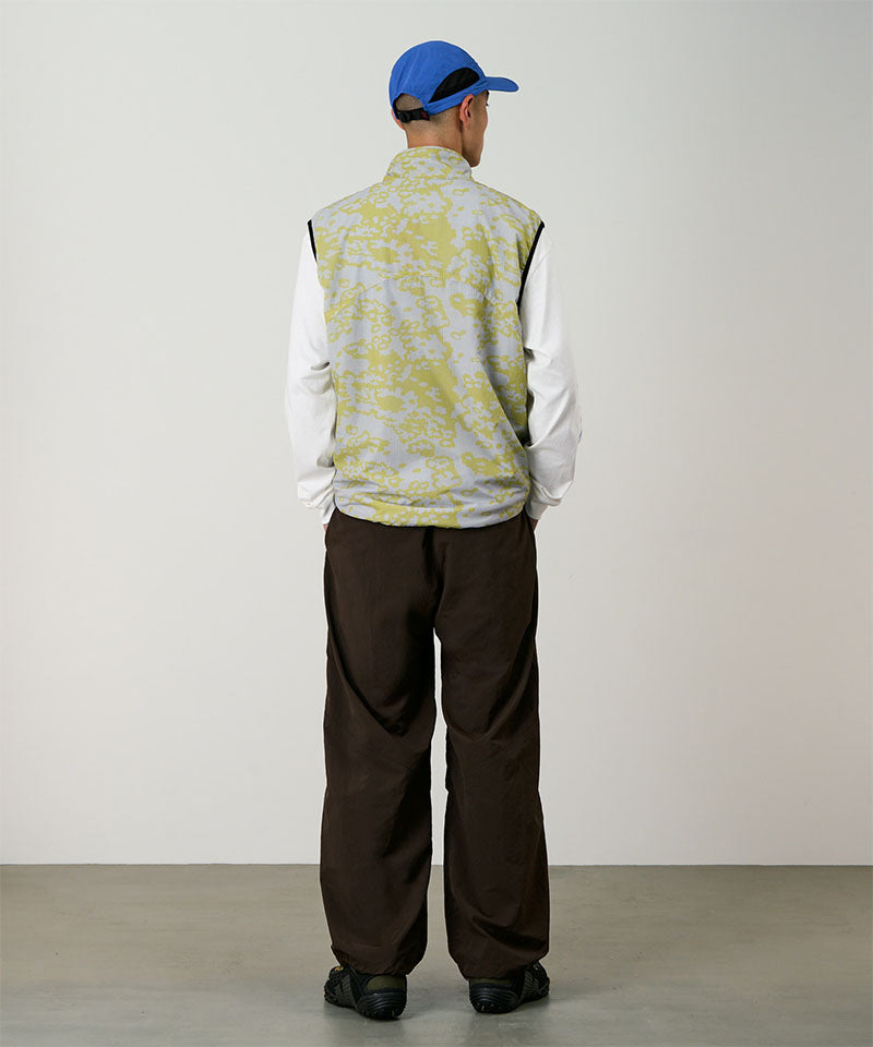 Back view of a model wearing a yellow and agrey grid camo Gramicci Flyweight Vest and brown pants on a light-tone background.